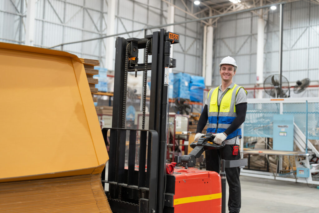 A ride-on powered pallet forklift being controlled by a man wearing a high-visibility jacket. 